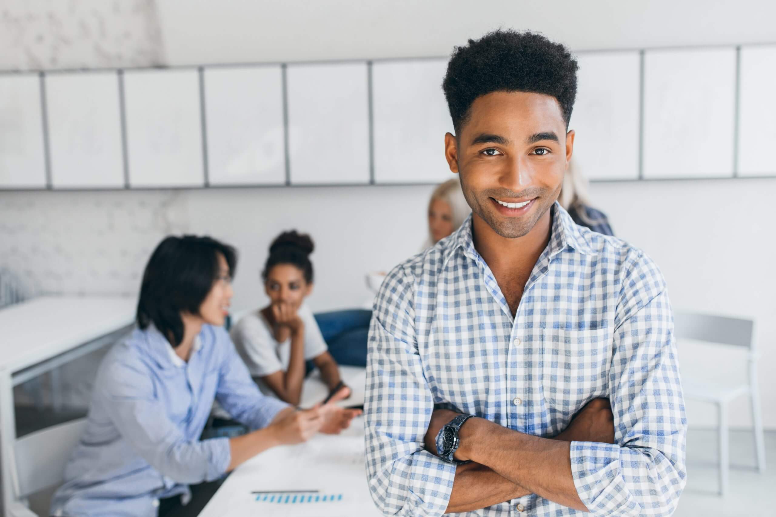 Glad young man with african hairstyle posing with arms crossed in his office with other employees on background. Male manager in blue shirt smiling during conference at workplace..